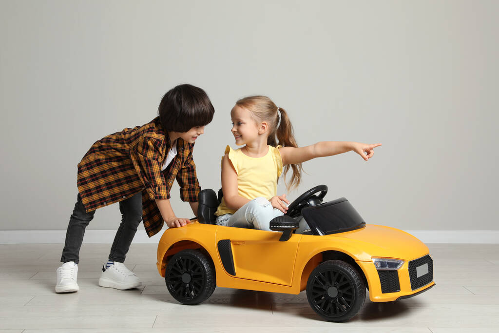 Симпатичний хлопчик штовхає дитячий електричний іграшковий автомобіль з маленькою дівчинкою біля сірої стіни в приміщенні
 - Фото, зображення