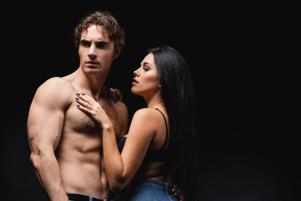 young woman in bra and jeans touching muscular boyfriend isolated on black  - Photo, Image