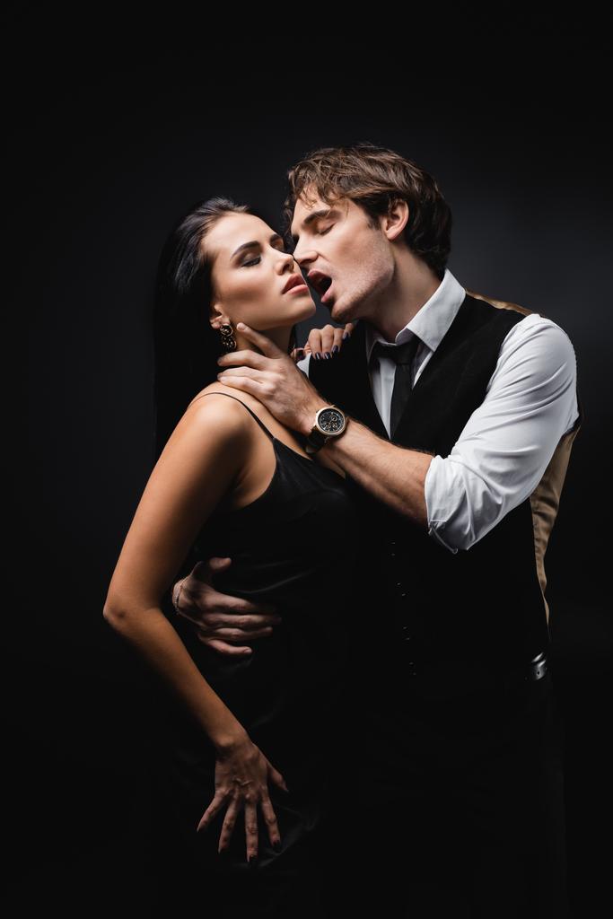 παθιασμένος άντρας με ανοιχτό στόμα αγκαλιάζει σαγηνευτική κοπέλα με ολισθηρό φόρεμα απομονωμένο σε μαύρο  - Φωτογραφία, εικόνα