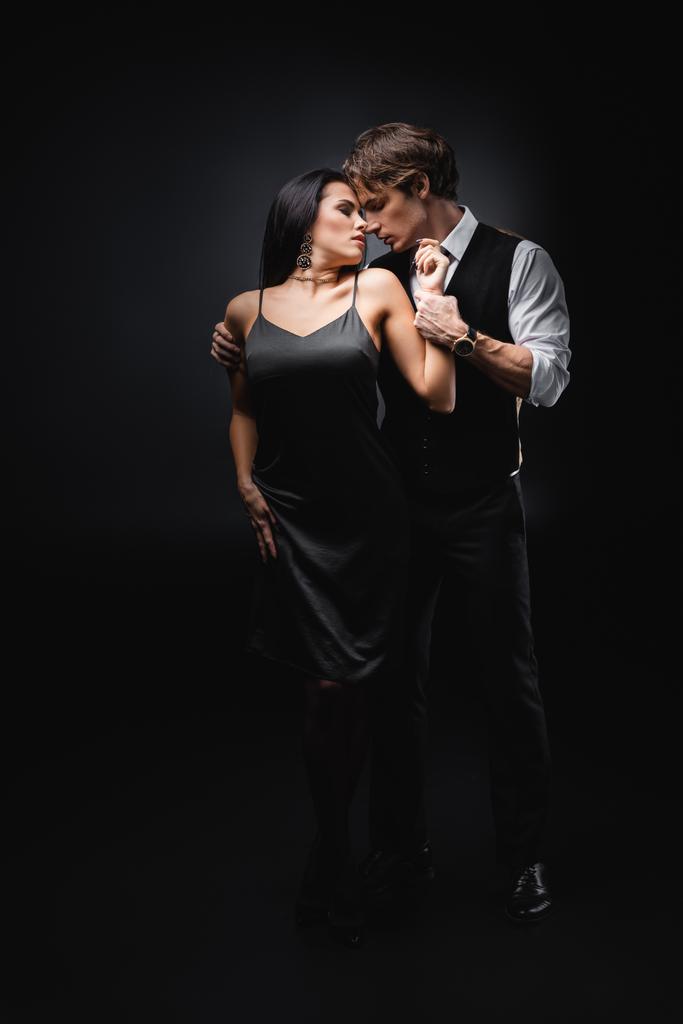 повна довжина чоловіка в жилеті і сорочці обіймає дівчину в сексуальній сукні ковзання на чорному
  - Фото, зображення