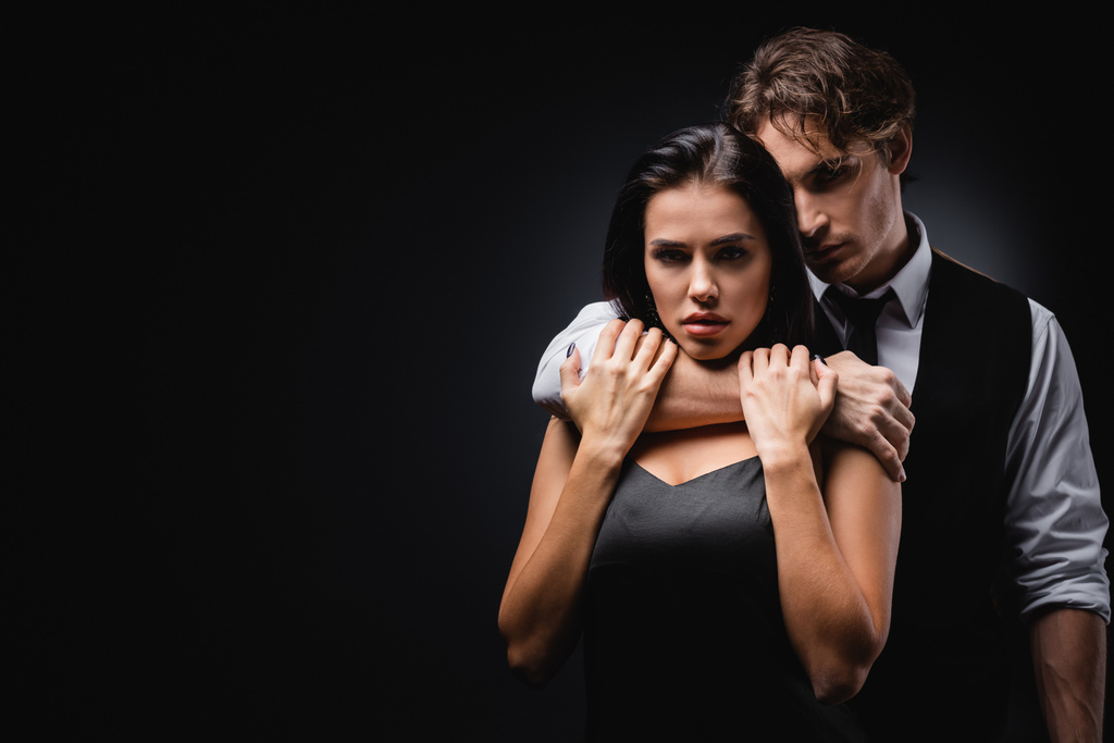 чоловік в жилеті і сорочці обіймає і задушує сексуальну дівчину в шовковій сукні ковзання ізольовано на чорному
 - Фото, зображення