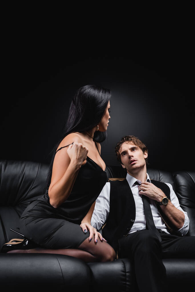 brunette femme ajustement sangle sur soie robe près passionné copain assis sur canapé sur noir  - Photo, image