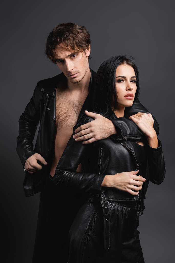 young man hugging brunette woman in black leather jacket isolated on grey - Photo, Image