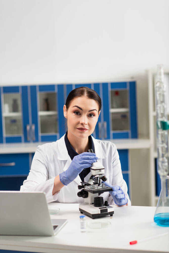 Brunetka naukowiec w rękawiczkach lateksowych patrząc na kamerę w pobliżu mikroskopu i laptopa w laboratorium  - Zdjęcie, obraz