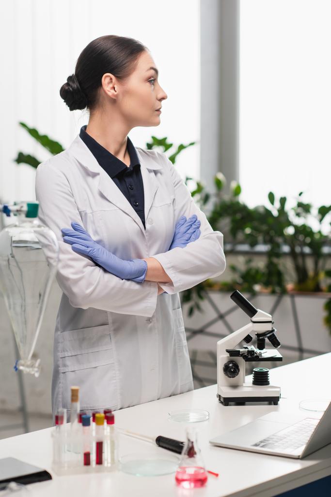 Вид сбоку на брюнетку ученого в белом халате, стоящего рядом с микроскопом и пробирками в лаборатории  - Фото, изображение