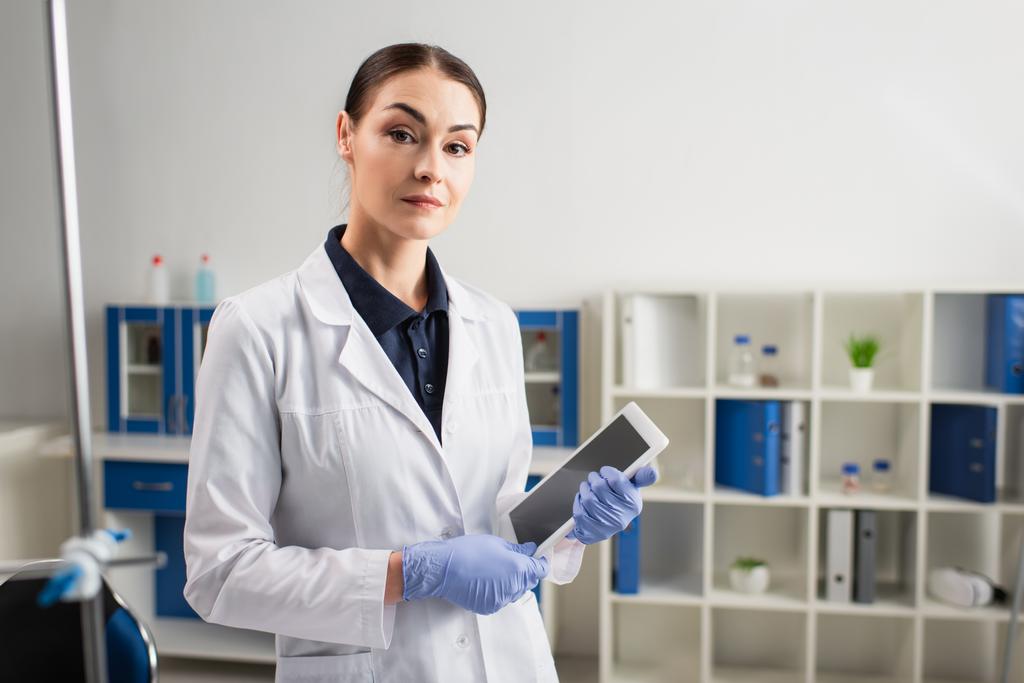 Ученый в латексных перчатках смотрит в камеру и держит цифровой планшет в лаборатории  - Фото, изображение