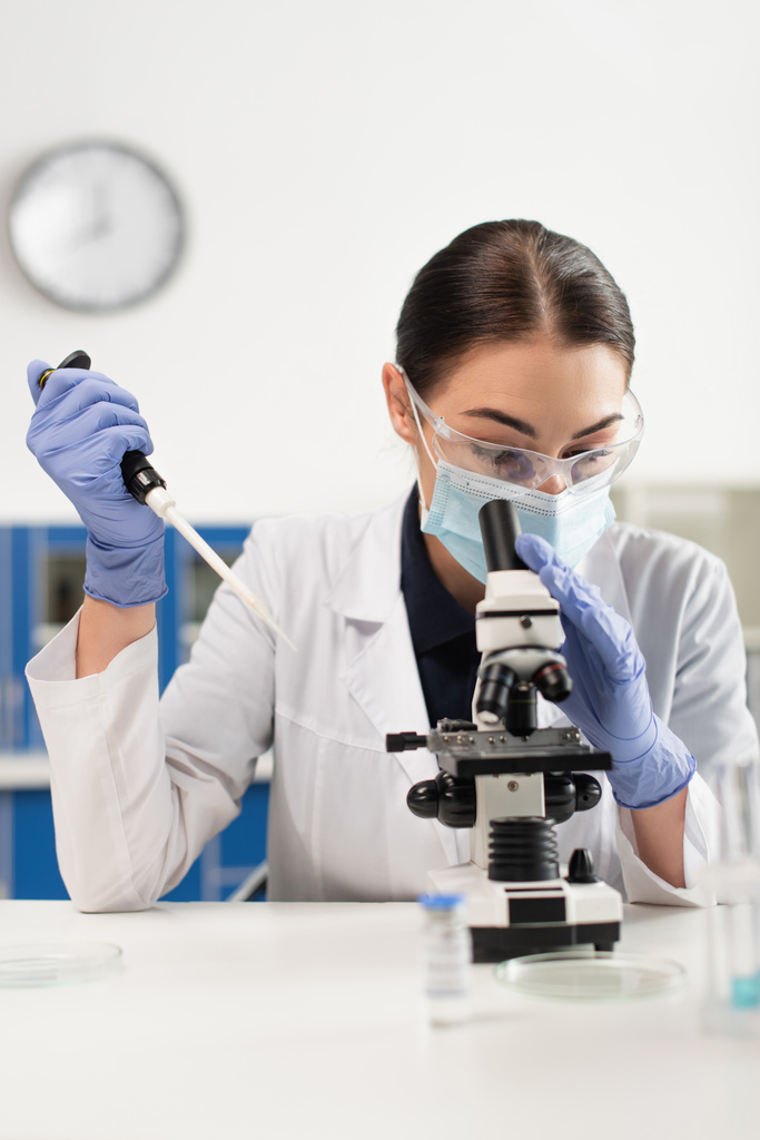 Tudós orvosi maszkban és latex kesztyűben, elektronikus pipettát tart a mikroszkóp és petri edények közelében a laborban  - Fotó, kép