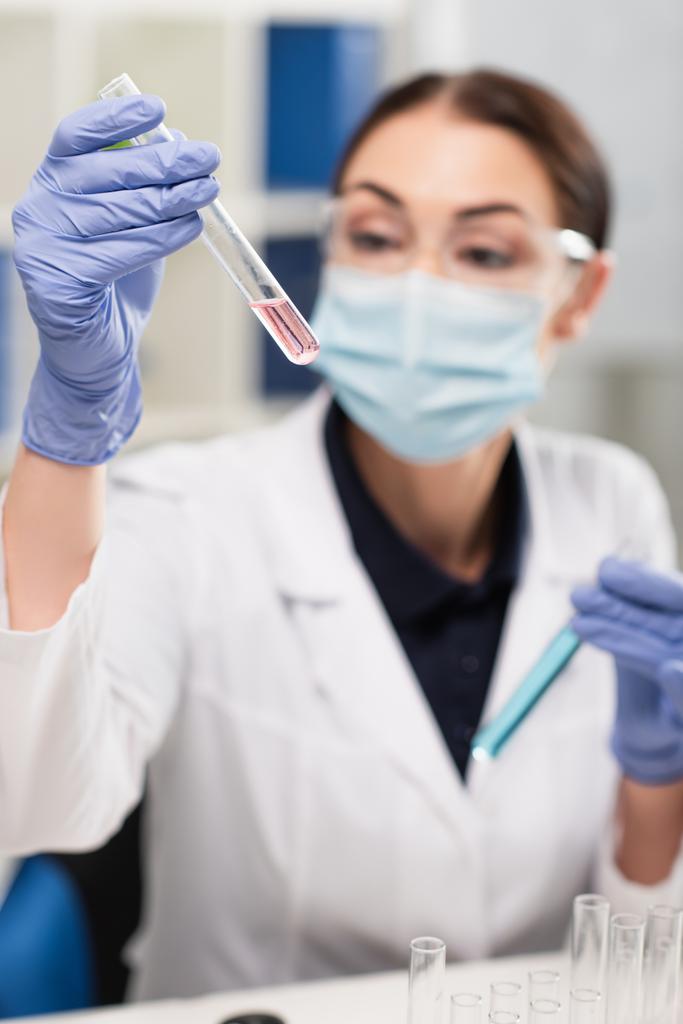 Tıbbi maskeli ve lateks eldivenli bulanık bilim adamı laboratuardaki test tüpüne bakıyor.  - Fotoğraf, Görsel