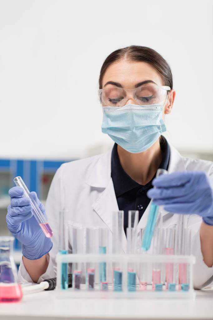 Ученый в латексных перчатках и медицинской маске держит пробирки возле пипетки и фляжки в лаборатории  - Фото, изображение