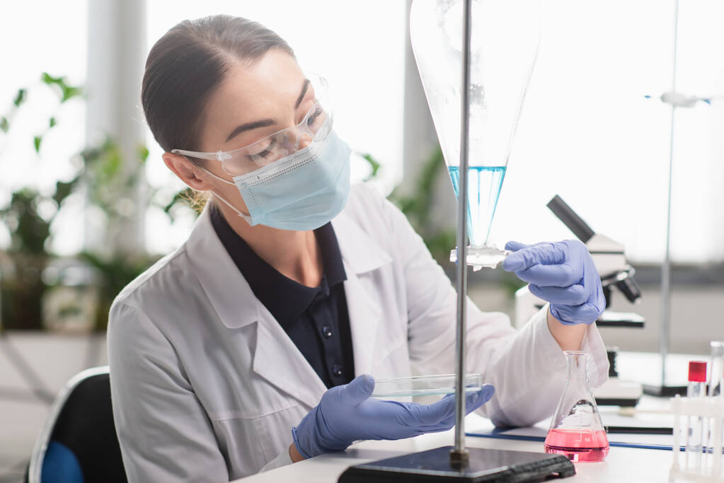 Brunette scientist in goggles and medical mask holding petri dish near flasks in lab  - Photo, Image