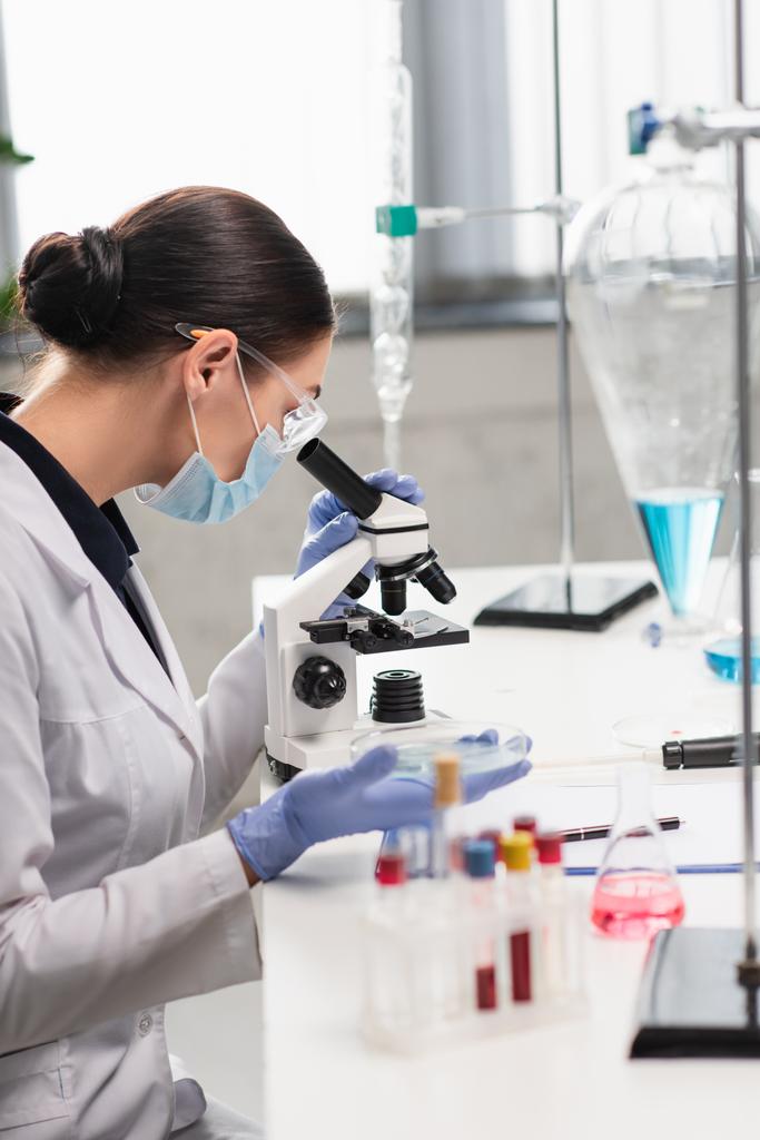 Scientifique en lunettes et masque médical utilisant un microscope près des fioles et des éprouvettes avec des échantillons de sang en laboratoire  - Photo, image