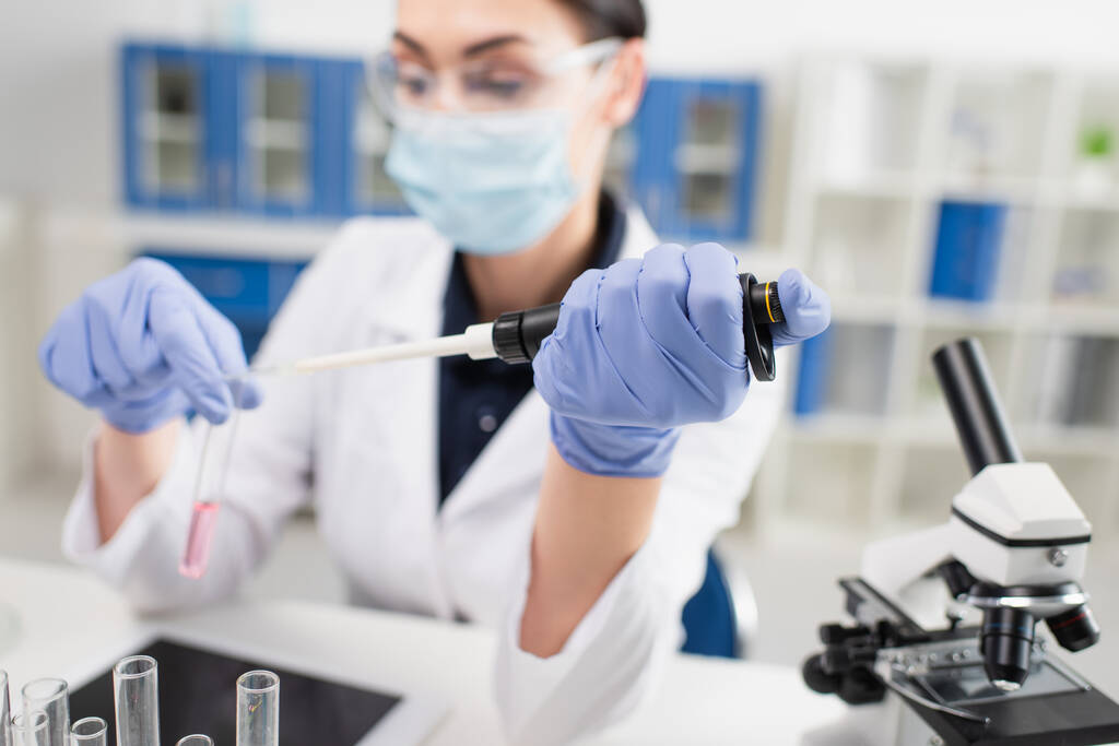Homályos tudós orvosi maszk kezében elektronikus pipetta közelében kémcsövek, mikroszkóp és digitális tabletta  - Fotó, kép