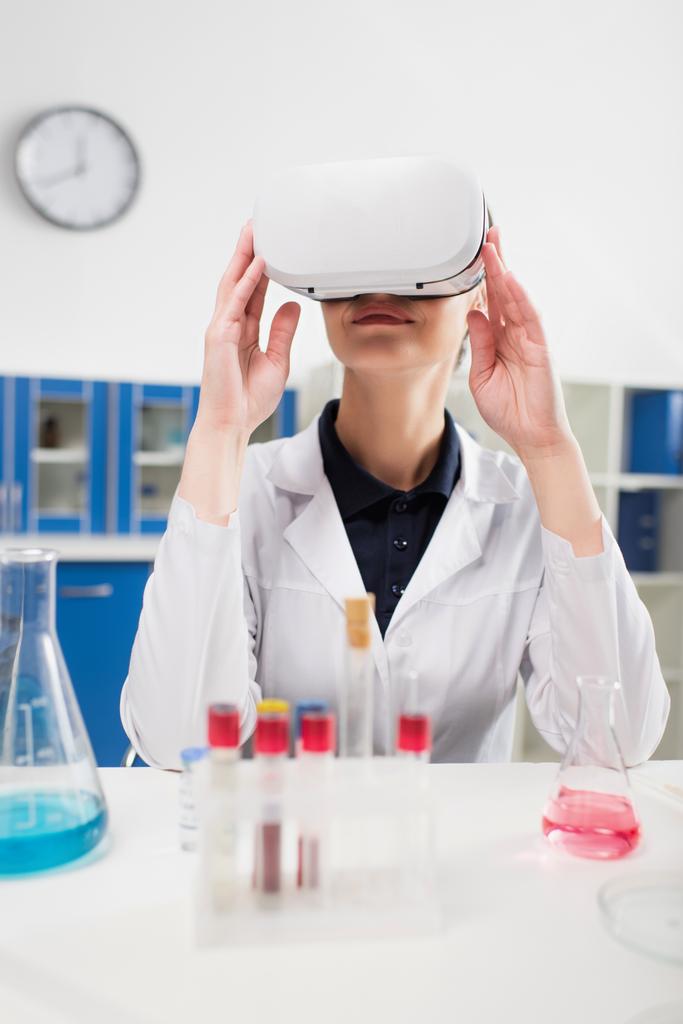 Un scientifique souriant dans un casque de réalité virtuelle près d'éprouvettes et de fioles floues en laboratoire  - Photo, image
