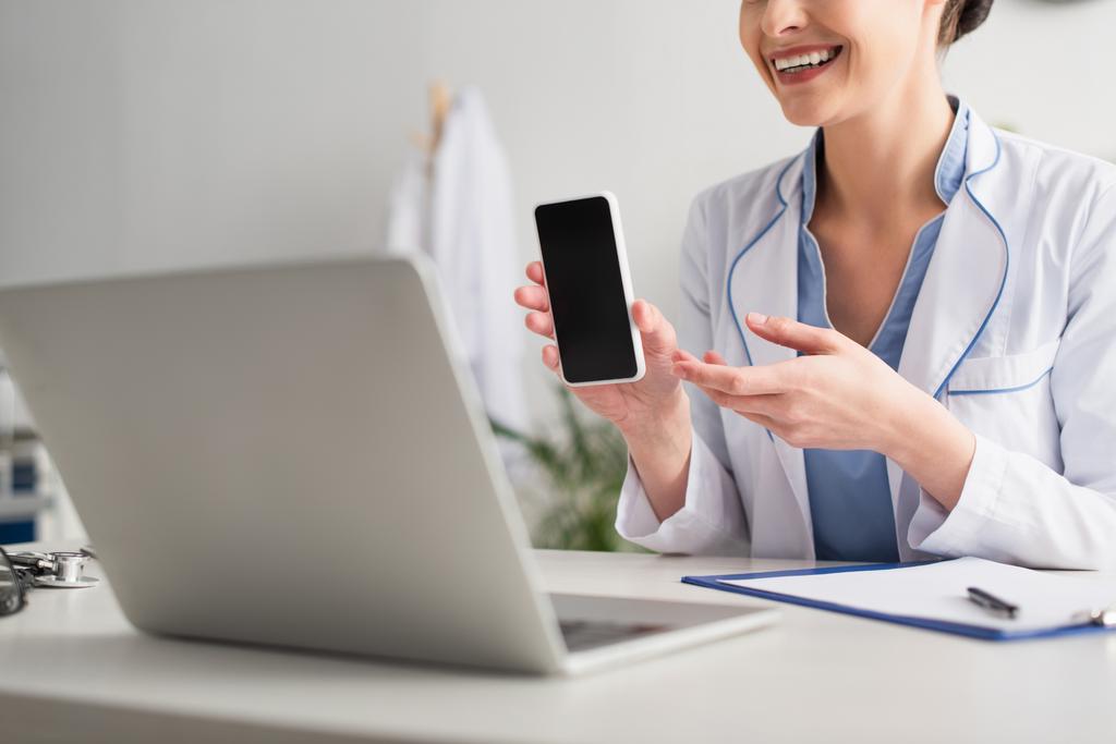 Vista cortada de sorrir médico segurando smartphone durante a consulta de vídeo no laptop na clínica  - Foto, Imagem