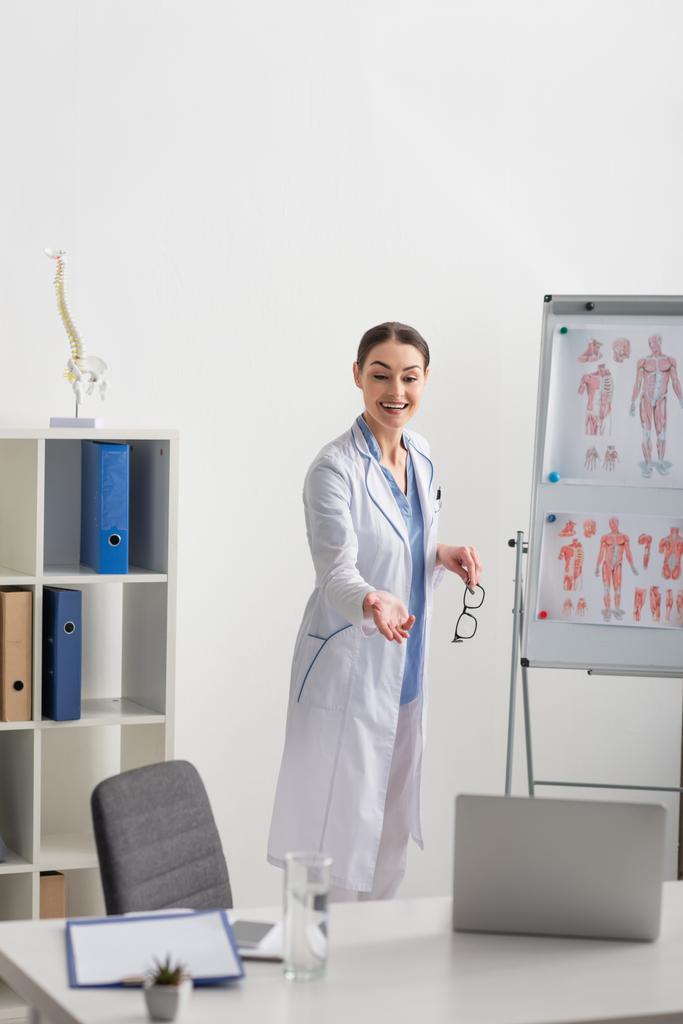 médico sorrindo apontando com a mão durante a chamada de vídeo, enquanto está perto de flip chart com imagens de anatomia - Foto, Imagem