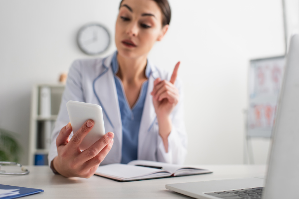 blurred doctor pointing with pen while looking at mobile phone at workplace - Photo, Image