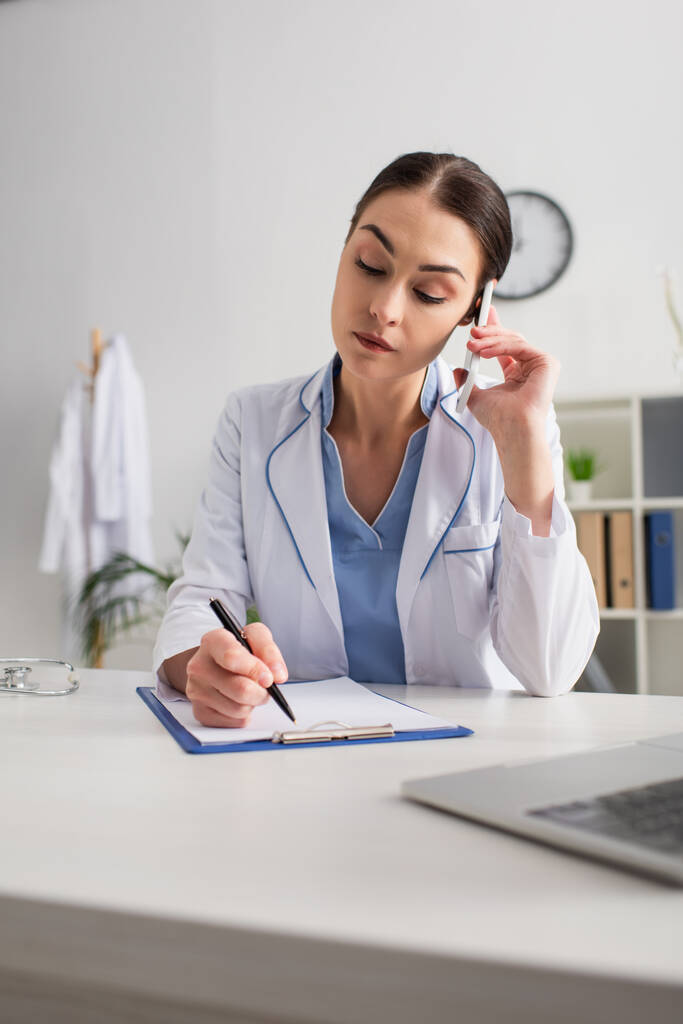 doctor talking on mobile phone while writing on clipboard near blurred laptop - Photo, Image