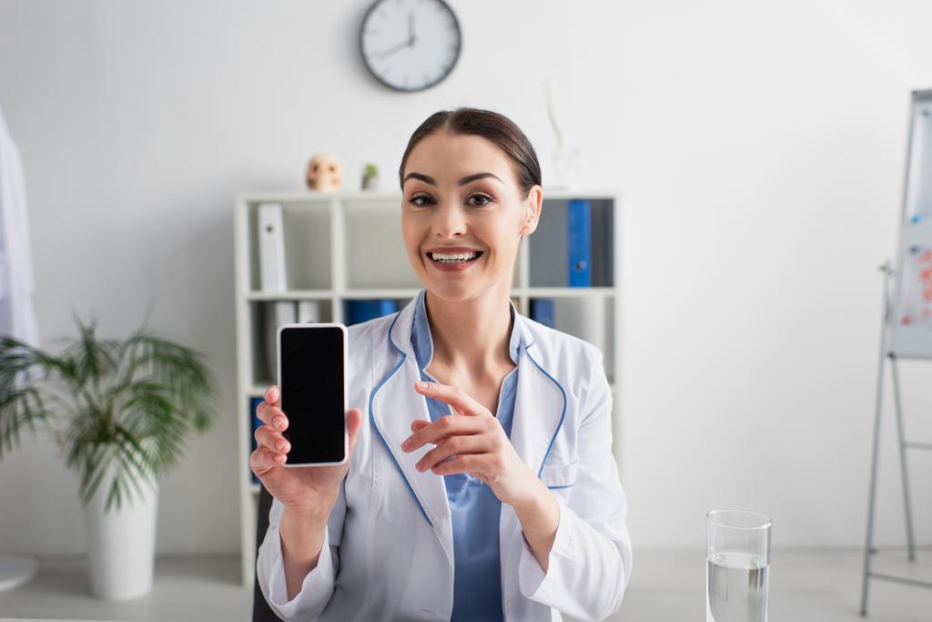 šťastný brunetka lékař ukazuje na mobilní telefon s prázdnou obrazovkou při pohledu na kameru - Fotografie, Obrázek