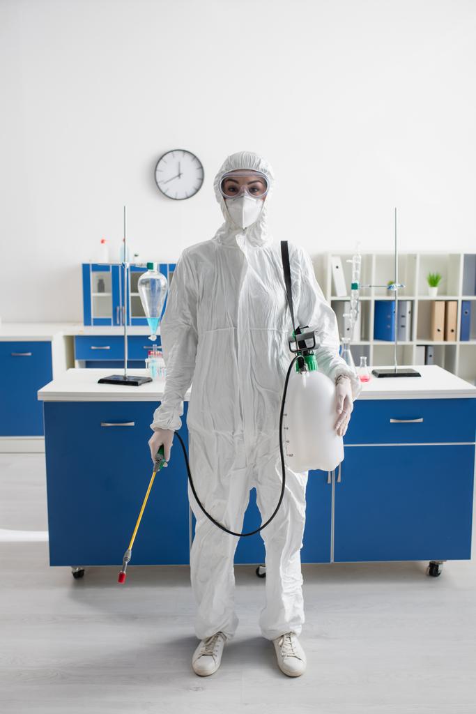 científico en traje de materiales peligrosos mirando a la cámara mientras sostiene el pulverizador en el laboratorio - Foto, imagen