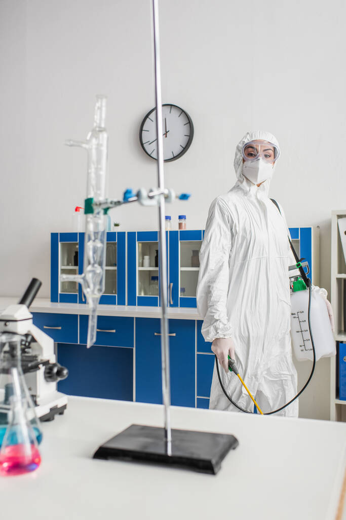 lekarz w kombinezonie ochronnym trzymający opryskiwacz patrząc na kamerę w laboratorium - Zdjęcie, obraz