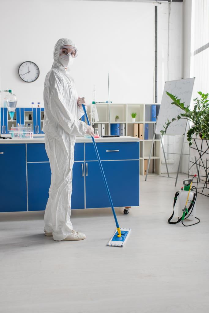モップで実験室の床を拭きながらカメラを見るハズマットスーツの科学者 - 写真・画像
