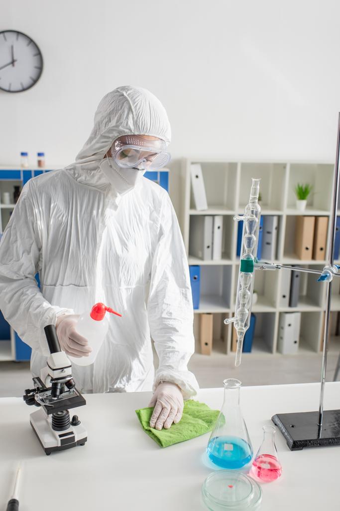 wetenschapper in hazmat pak afvegen bureau in lab in de buurt van kolven en microscoop - Foto, afbeelding