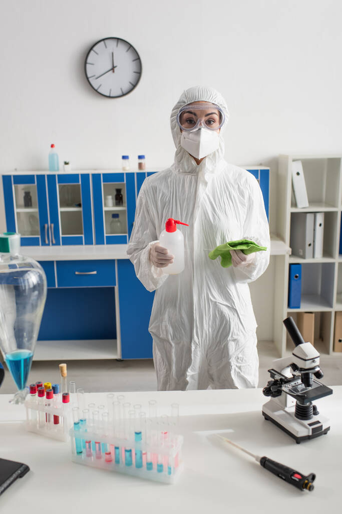 naukowiec w kombinezonie ochronnym trzymający butelkę z detergentem i szmatą patrząc w kamerę  - Zdjęcie, obraz