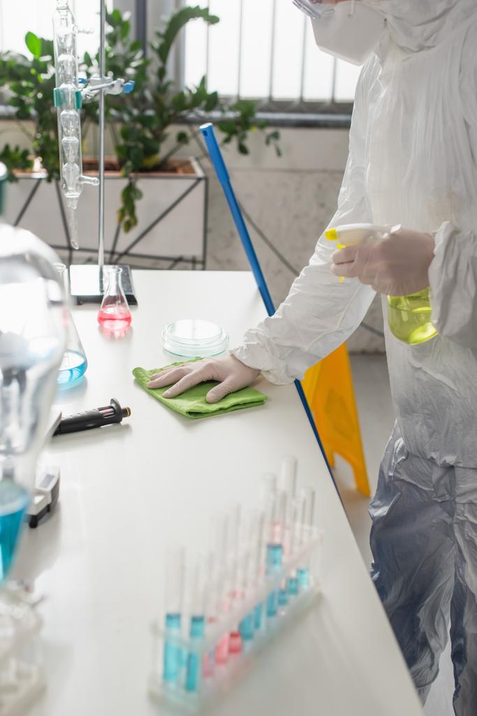 científico en traje protector que limpia el escritorio con el trapo mientras sostiene la botella del aerosol - Foto, imagen