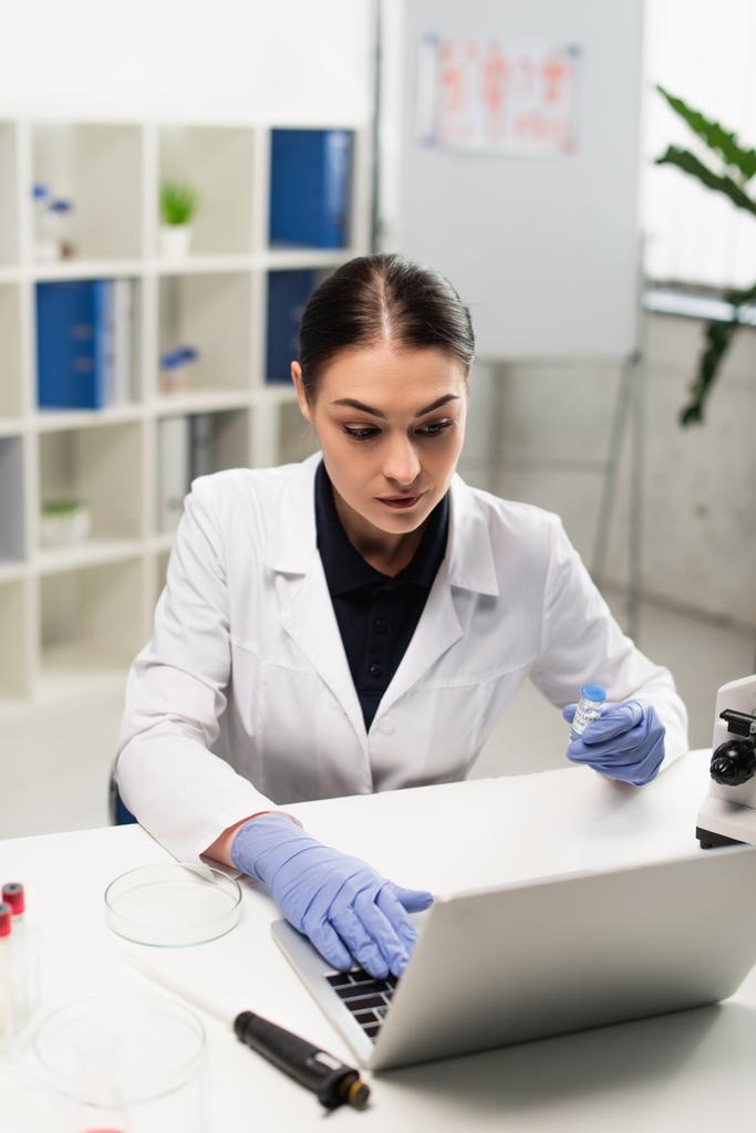 Brünette Wissenschaftlerin hält Impfstoff in der Hand, während sie auf Laptop in der Nähe der Petrischale tippt - Foto, Bild