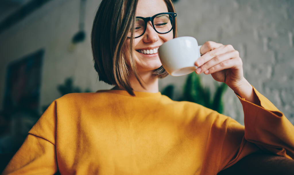 Zębowa hipsterka w okularach optycznych dla zapewnienia korekcji wzroku ciesząca się porannym czasem picia smacznego napoju kofeinowego w lokalnej stołówce, pogodna kobieta trzymająca kubek kawy i uśmiechnięta - Zdjęcie, obraz