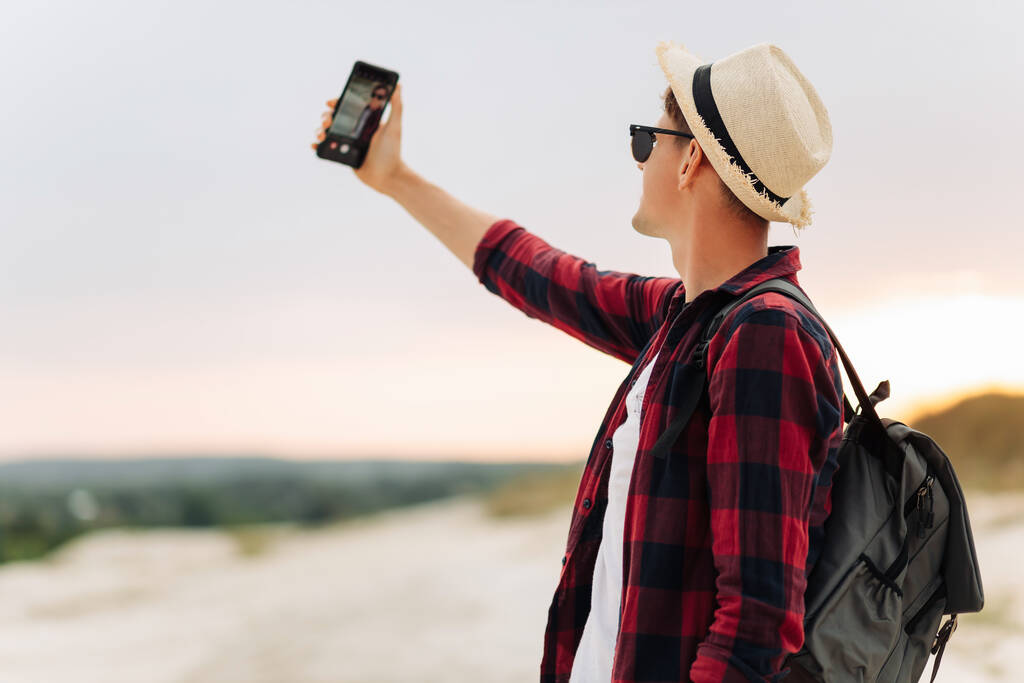 Młody turysta w okularach przeciwsłonecznych i kapeluszu robi sobie selfie podczas spaceru po lesie. Podróżnik prowadzący zdrowy aktywny tryb życia, cieszący się przyrodą, spacerujący w przyrodzie, letni spacer w górach - Zdjęcie, obraz