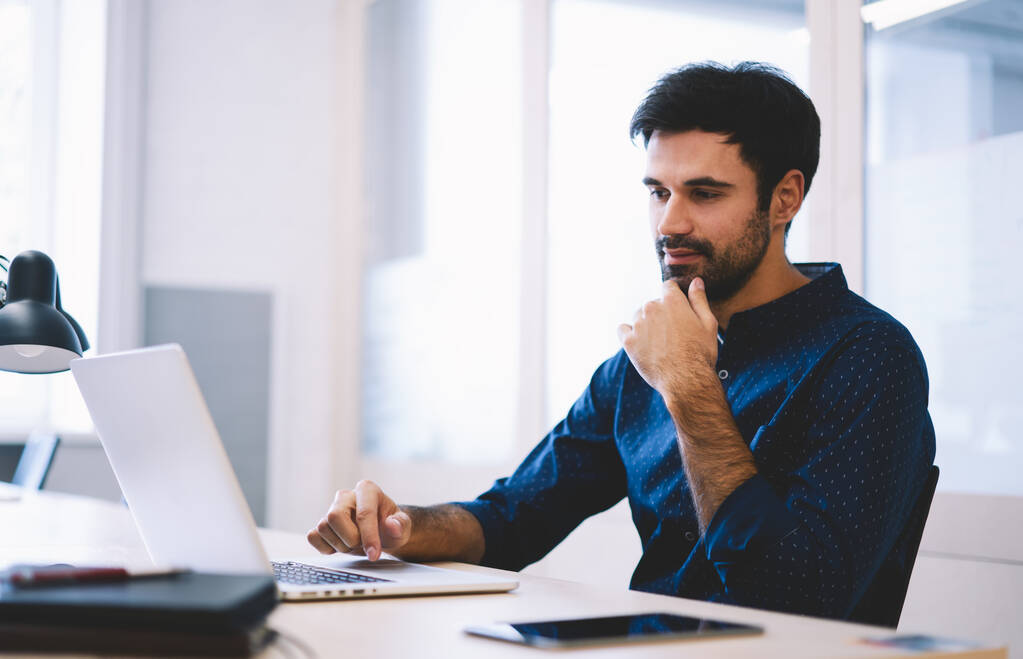 Soustředěný vousatý muž v košili dotýkající se brady sedící u stolu s laptopem při práci na obchodním projektu na lehkém moderním pracovišti - Fotografie, Obrázek