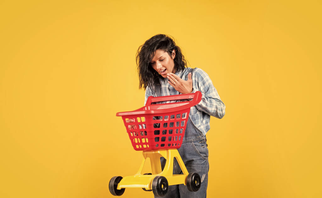 iloinen nainen paita denim tyyli kuljettaa ostoskärry keltaisella pohjalla, ostoksia - Valokuva, kuva