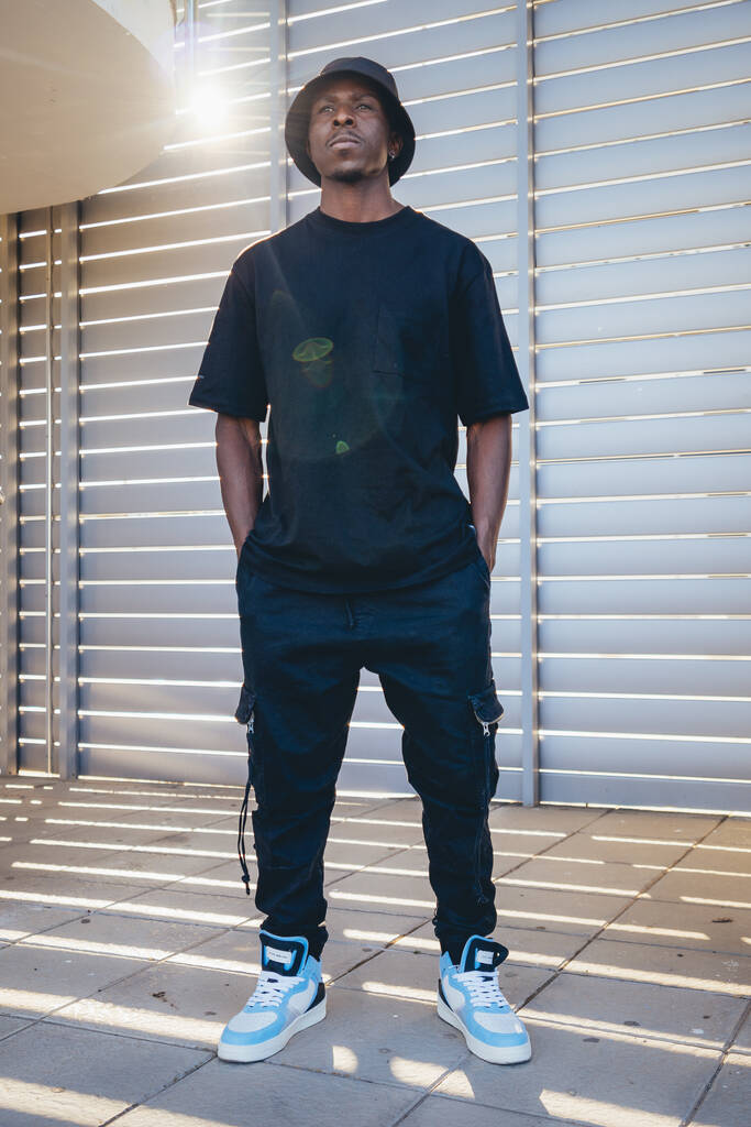 Plan vertical d'un jeune homme noir vêtu de vêtements de rue debout derrière des volets par une journée ensoleillée - Photo, image