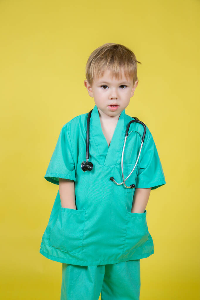 Doktor kılığına girmiş küçük beyaz bir çocuğun portresi. Sarı üzerine izole edilmiş yeşil ceket. - Fotoğraf, Görsel