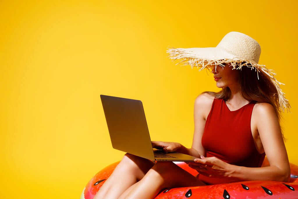 La mujer joven en la paja se sienta en el anillo inflable trabaja en el ordenador portátil sobre el fondo amarillo. independiente. Viajes - Foto, imagen