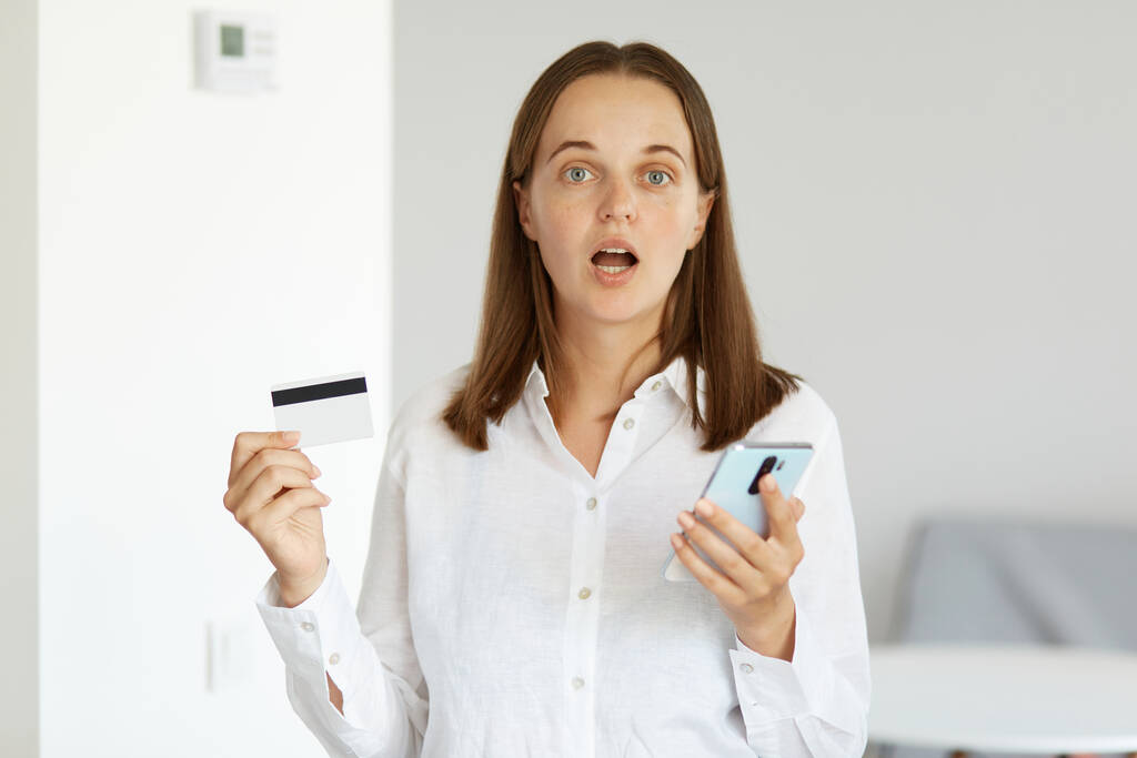 Zszokowana kobieta w białej koszuli stojąca w pomieszczeniu z kartą kredytową i telefonem komórkowym w rękach, zaskoczona wygraną na loterii lub zakładami. - Zdjęcie, obraz