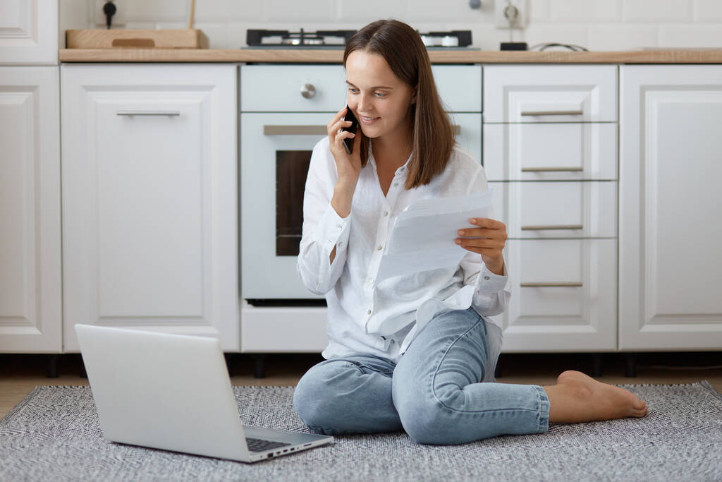 Indoor shot of smiling attractive young adult woman sitting on floor in kitchen and using laptop and smart phone, talking with client, having business online, holding documents in hands. - Foto, Imagen