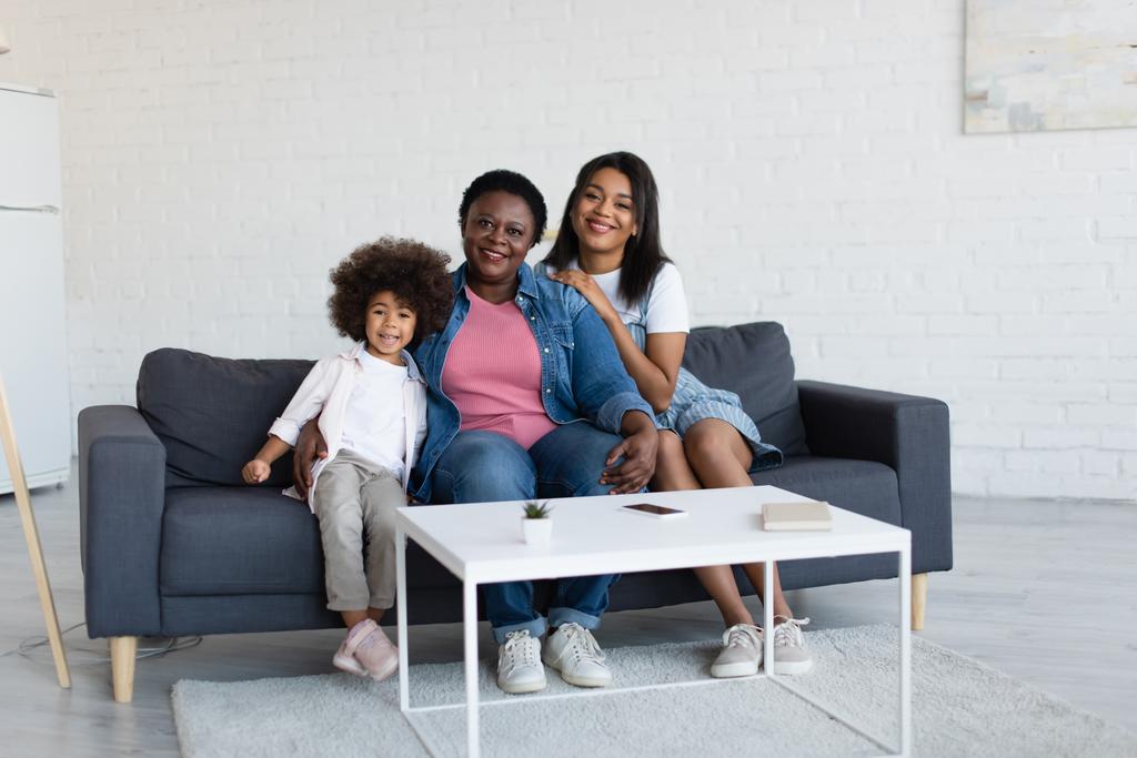 iloinen afrikkalainen amerikkalainen tyttö äiti ja mummo katselee kameraa istuessaan sohvalla - Valokuva, kuva