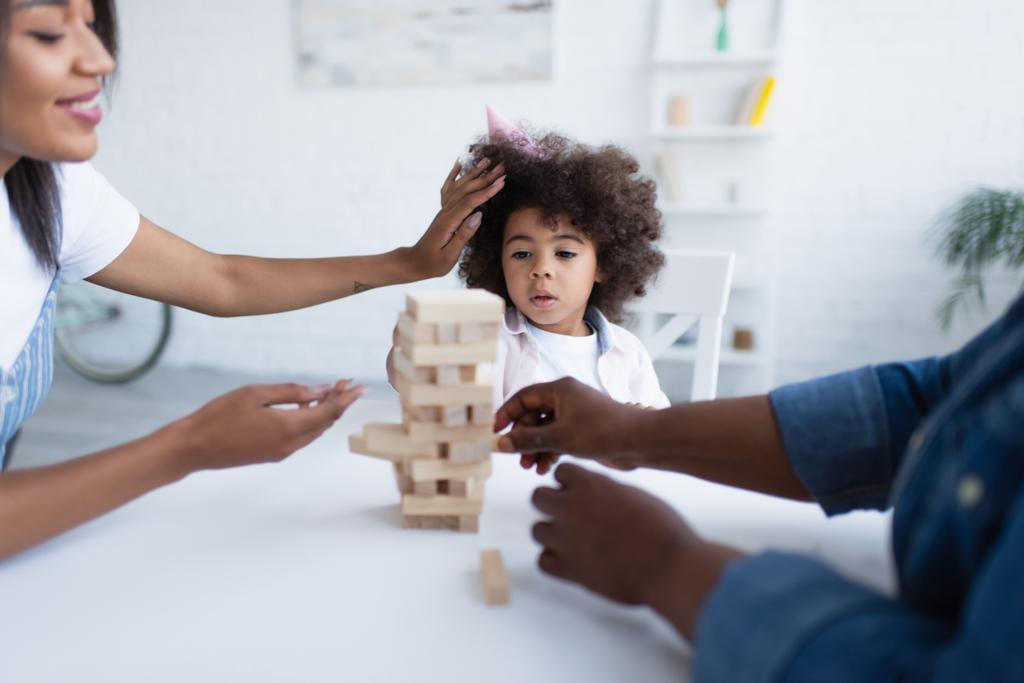 african american girl in party cap playing wood blocks game with blurred mom and granny - Photo, Image