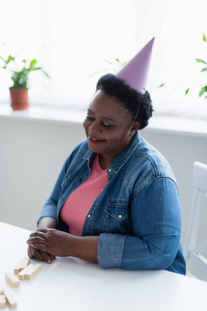 donna afroamericana di mezza età in berretto da festa sorridente mentre sedeva vicino a blocchi di legno gioco sul tavolo - Foto, immagini