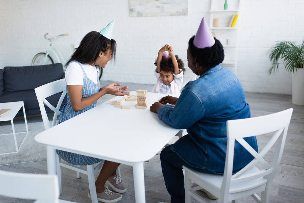 Afryki amerykanka w party cap wskazując na drewnianą wieżę na stole w pobliżu szczęśliwej córki - Zdjęcie, obraz