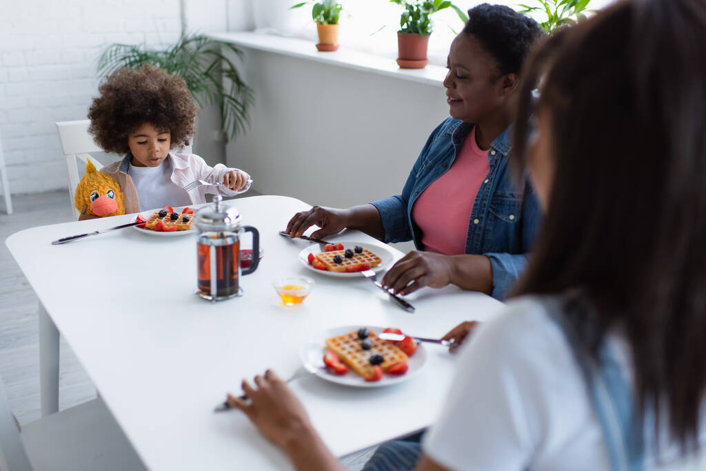 アフリカ系アメリカ人の女の子は柔らかいおもちゃでテーブルに座っている間におばあちゃんとぼやけたお母さんと朝食を持っています - 写真・画像