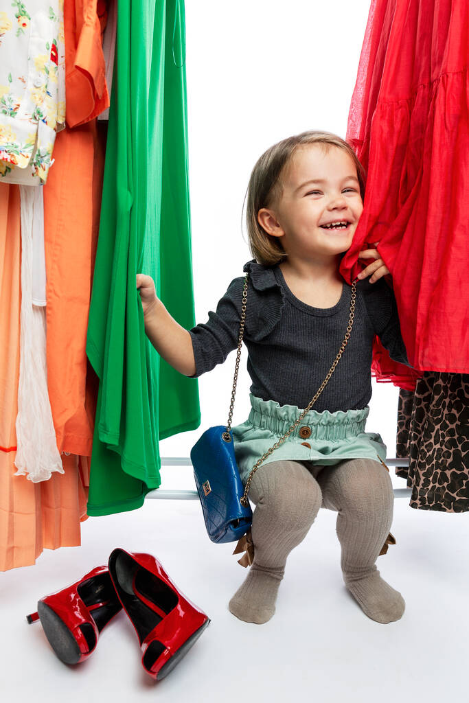 Petite fille souriante en chaussures rouges de maman parmi les robes sur cintres. Délice, bonheur et mode. Vertical. - Photo, image