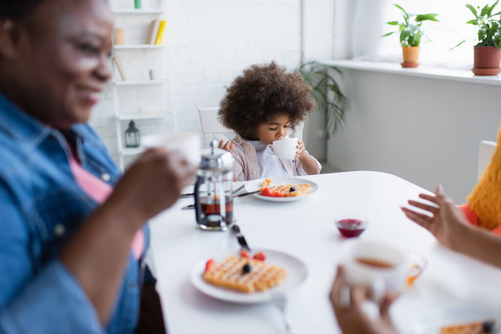 Afrykańskie amerykańskie dziecko pijące herbatę podczas śniadania z rozmazaną babcią i mamą  - Zdjęcie, obraz