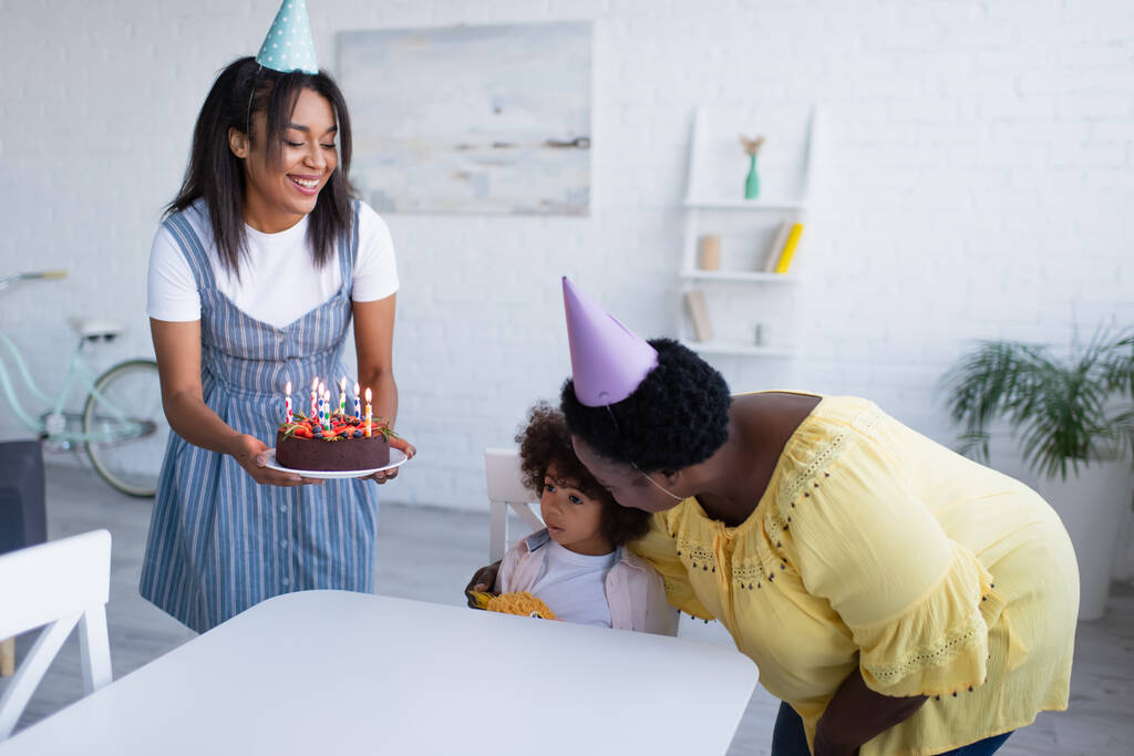 Afrikanerin sitzt am Tisch neben Oma und umarmt sie und Mutter mit Geburtstagstorte - Foto, Bild