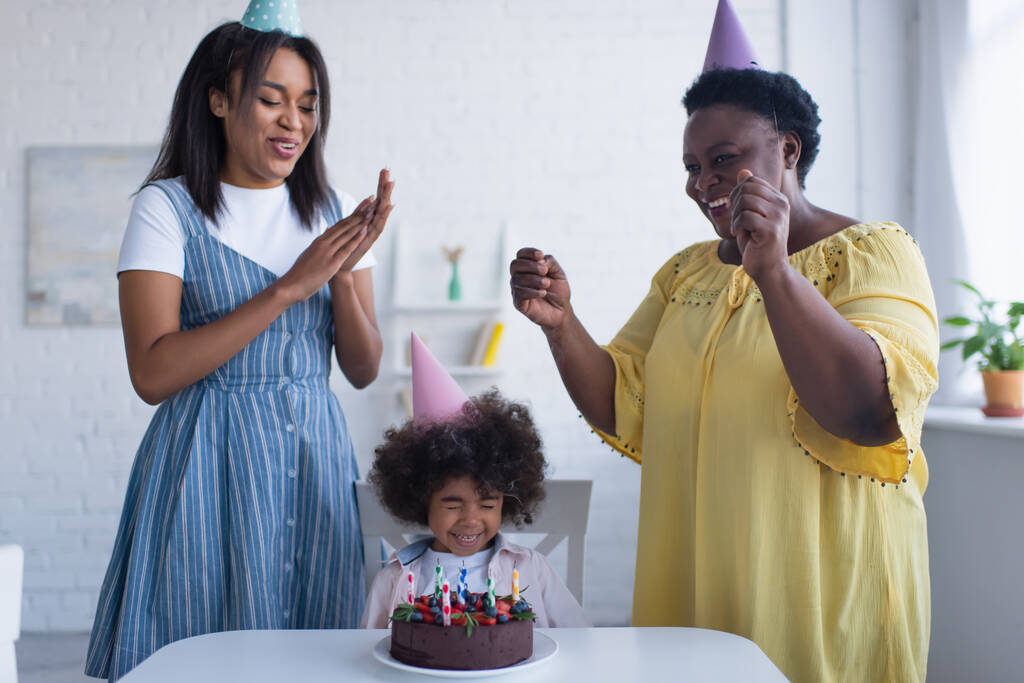 kisbaba afro-amerikai lány csukott szemmel ül közel születésnapi torta és anya nagymama party sapkás - Fotó, kép