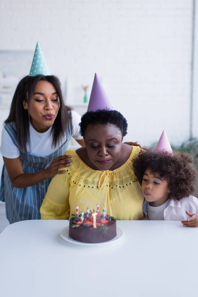 Afrykańskie amerykanki i dziewczynka w czapkach imprezowych zdmuchując świeczki na torcie urodzinowym - Zdjęcie, obraz