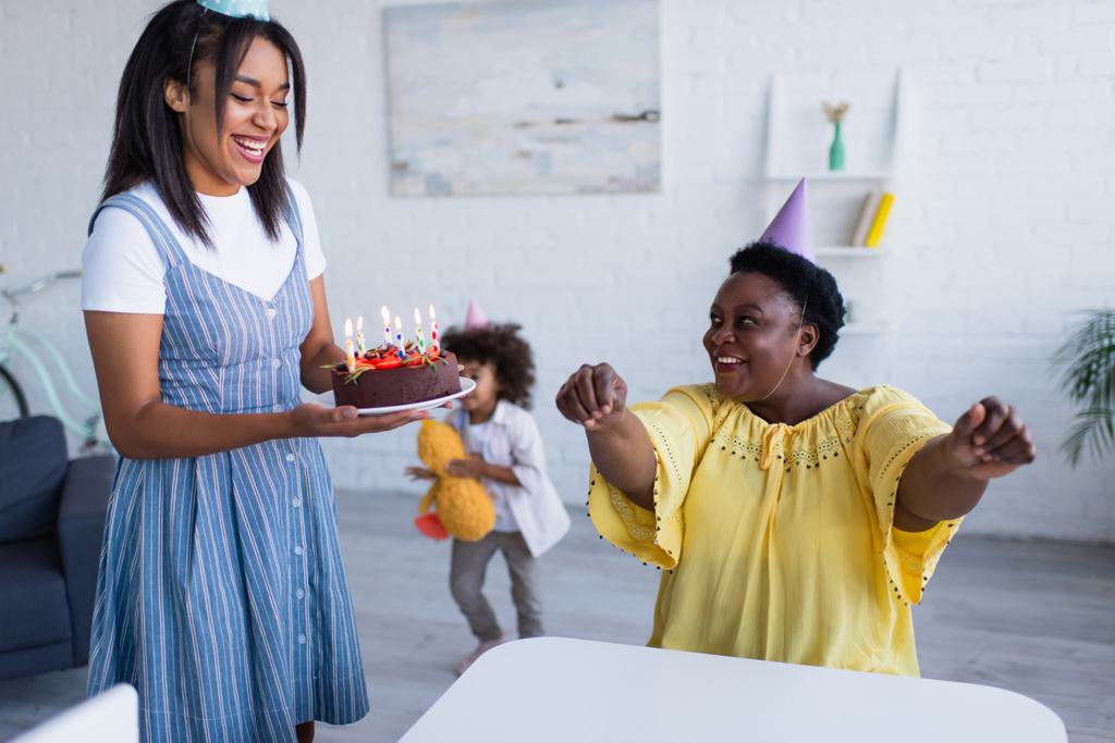 jovem afro-americana segurando bolo de aniversário perto de mãe feliz em boné de festa - Foto, Imagem