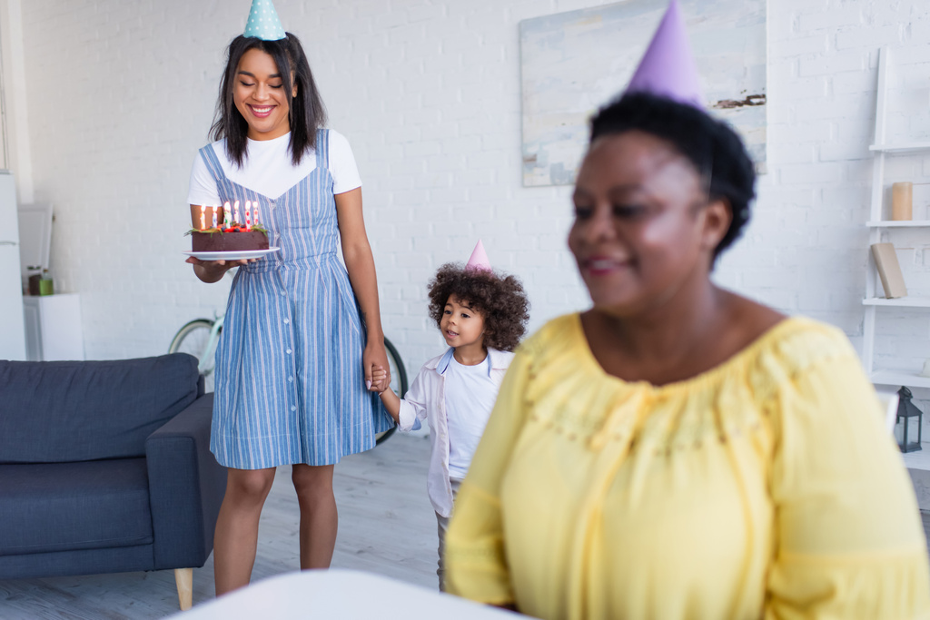smiling african american mother and daughter with birthday cake near blurred granny in party cap   - Photo, Image